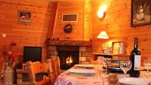 Un intérieur chaleureux en bois, avec une cheminée, une table à manger dressée pour deux personnes et un téléviseur fixé au mur.