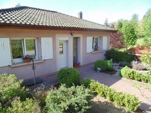 A house with a tiled roof, white shutters, and a front door. There are potted plants on the windowsills and a garden with various shrubs and a small tree. - Charming Mountain Cottage with Fireplace, Veranda, Garage, and WiFi in Peaceful Setting (Anould)