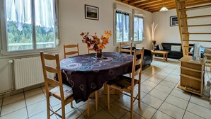 A dining area with a round table, chairs, a vase of flowers, and a view of the outdoors through large windows. - Nature Getaway: Cozy Gîte, Near Ski Trails & Lake, Gérardmer (Gérardmer)