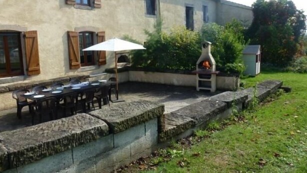 An outdoor patio with a stone table and chairs, a pizza oven, and a small shed in a garden setting.