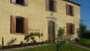 A two-story building with wooden shutters on the windows, a green sign indicating a guest room, and a small garden area with plants and a bench.