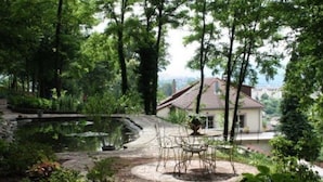Ein Haus, umgeben von einem Teich und üppiger Bepflanzung, mit einer Terrasse und Gartenmöbeln.