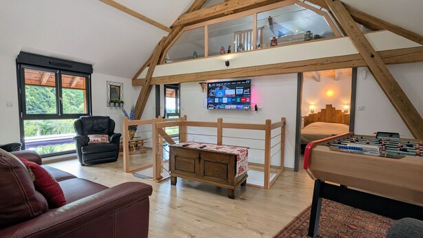 Een loft-achtige kamer met een houten plafond, een bank, een tafel met een tafelvoetbalspel en een televisie aan de muur.