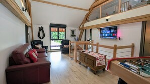 Een ruime kamer met een houten plafond, een bank, een stoel, een tafeltje met een tafelvoetbalspel en een aan de muur bevestigde televisie.