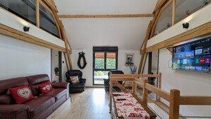 Een ruime woonkamer met een hoge nok, een houten trap, een leren bank en een aan de muur bevestigde televisie.