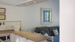 A modern kitchen with white cabinets, a microwave, and a white countertop. There is a grey sofa with a cactus pillow and a framed picture on the wall.