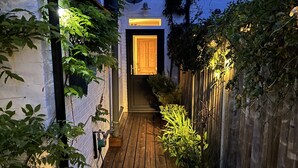 A narrow wooden pathway leading to a door with a window above, surrounded by greenery and a white brick wall. - Lilly Cottage - Fisherman's Cottage Whitstable (Whitstable)
