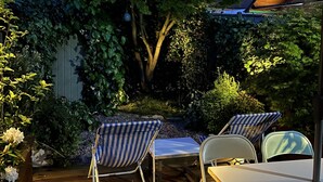 A wooden deck with two striped lounge chairs and a table, surrounded by lush greenery and a large umbrella. - Lilly Cottage - Fisherman's Cottage Whitstable (Whitstable)