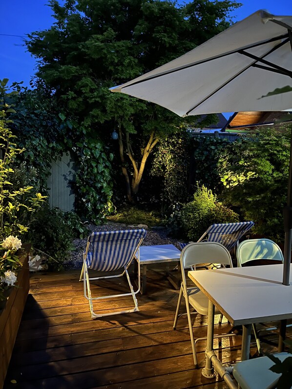 A wooden deck with two striped lounge chairs and a table, surrounded by lush greenery and a large umbrella. - Lilly Cottage - Fisherman's Cottage Whitstable (Whitstable)