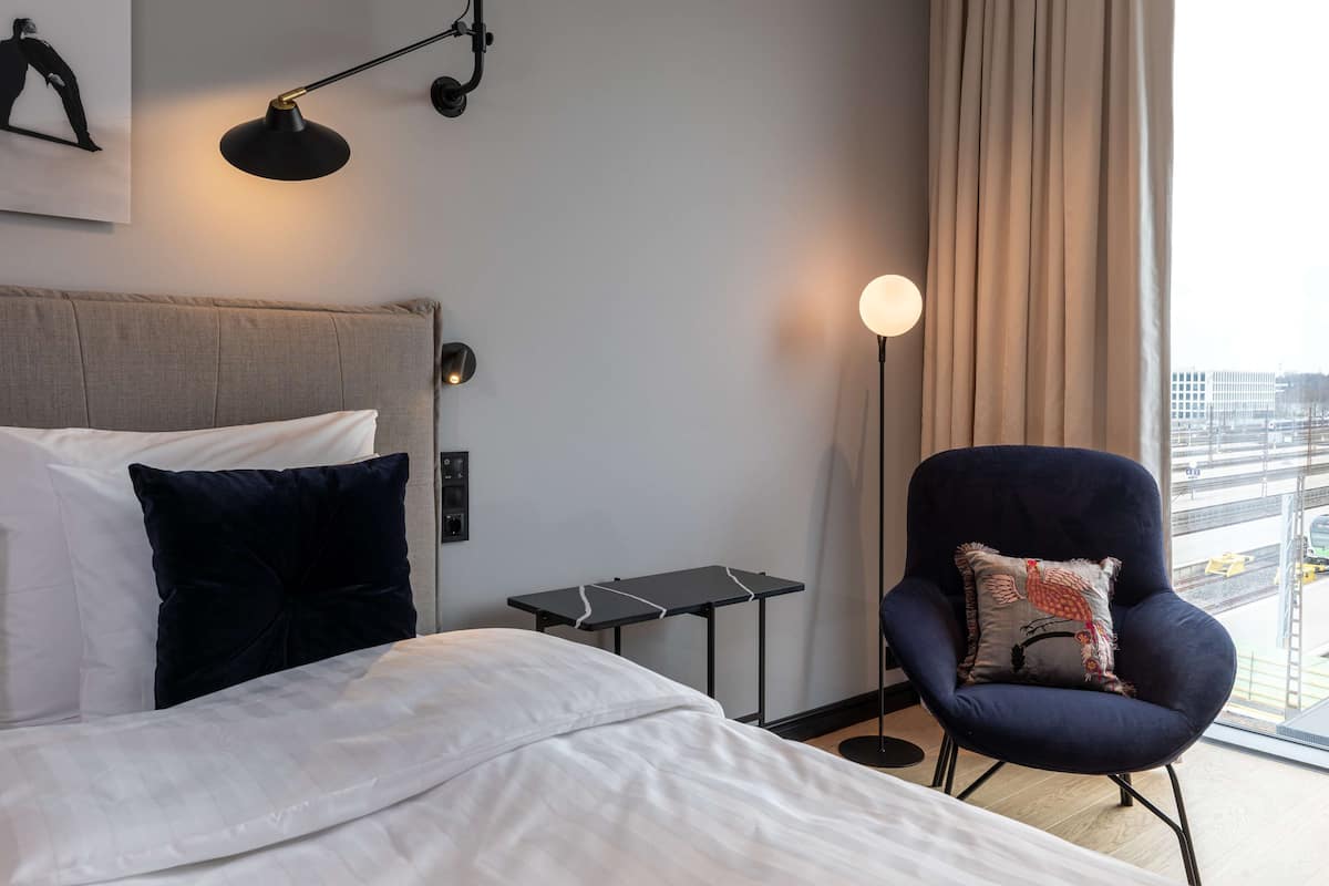 A hotel room with a bed, a chair, a small table, and a view of the city through the window.