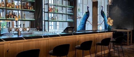 A bar area with wooden bar counter, black chairs, shelves with liquor bottles, glasses, and decorative peacock statues.