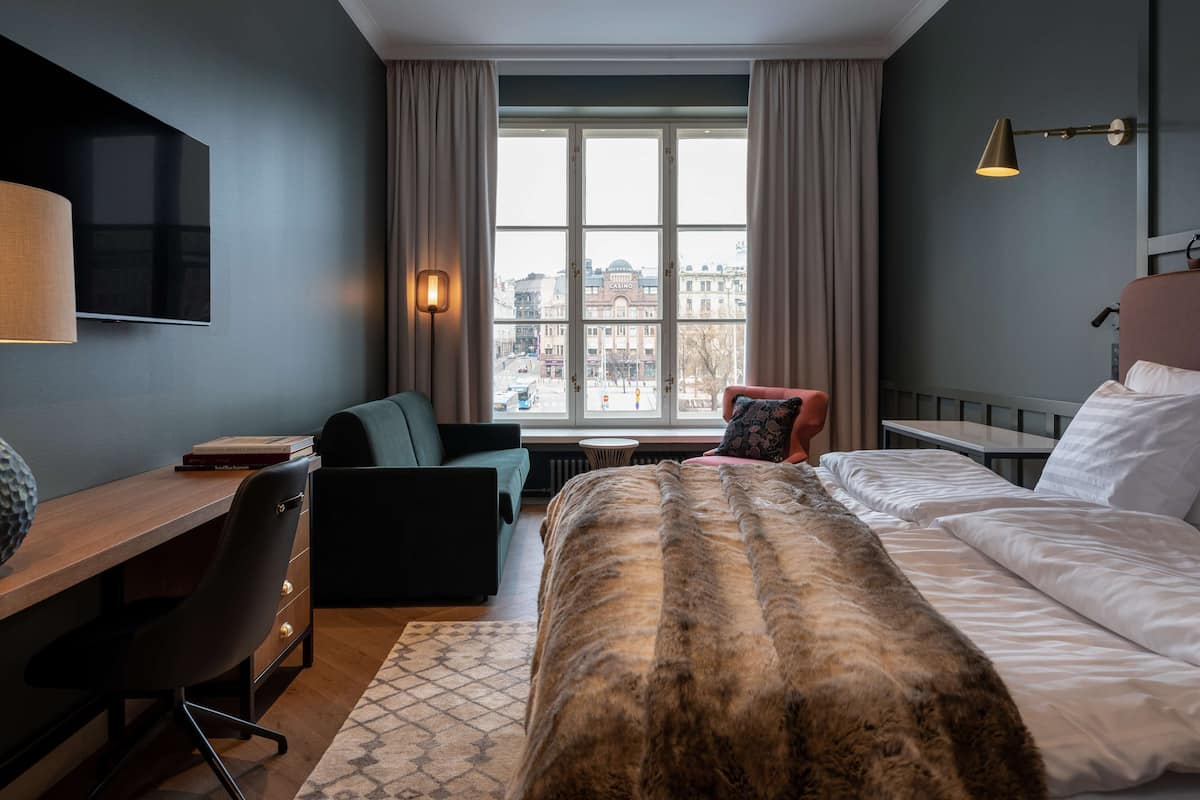 A modern hotel room with a large bed, a desk, a chair, and a view of the city through large windows.