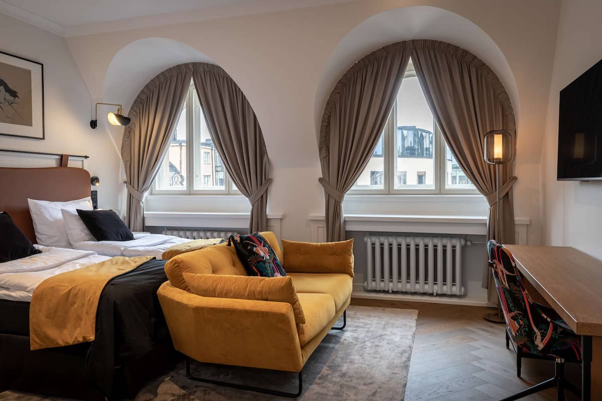 A hotel room with a bed, a sofa, a desk, and two windows with curtains.