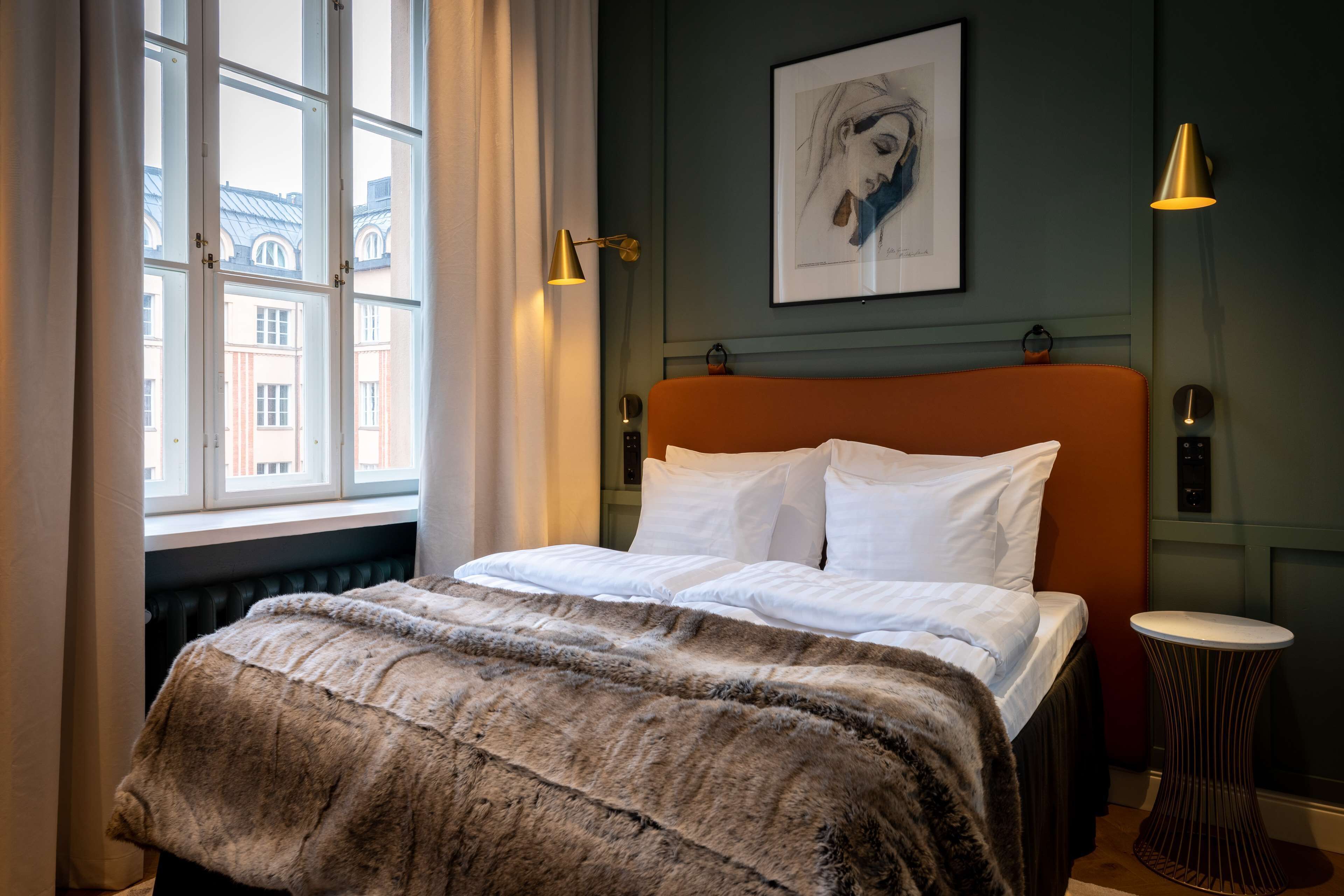 Habitación de hotel con cama, ventana con cortinas, una obra de arte enmarcada en la pared y una mesita de noche con lámpara.