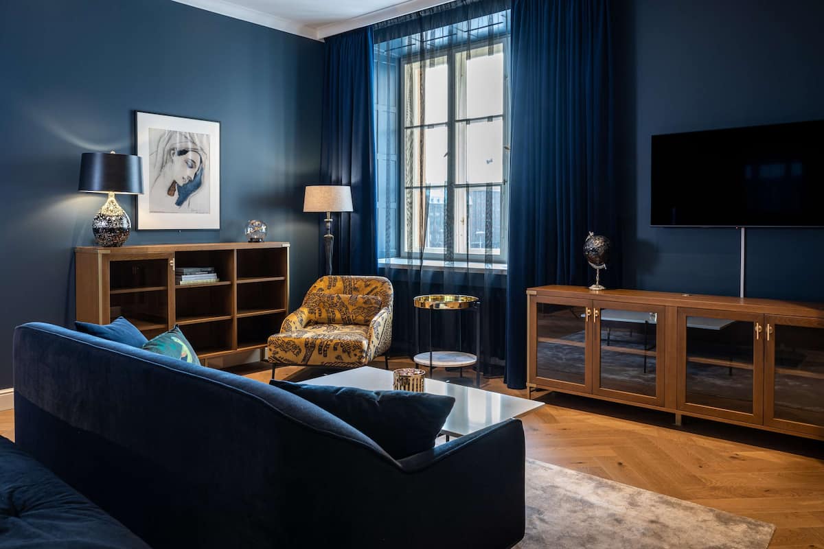 A modern living room with a dark blue sofa, a patterned armchair, a coffee table, a bookshelf, a TV, and a large window with curtains.