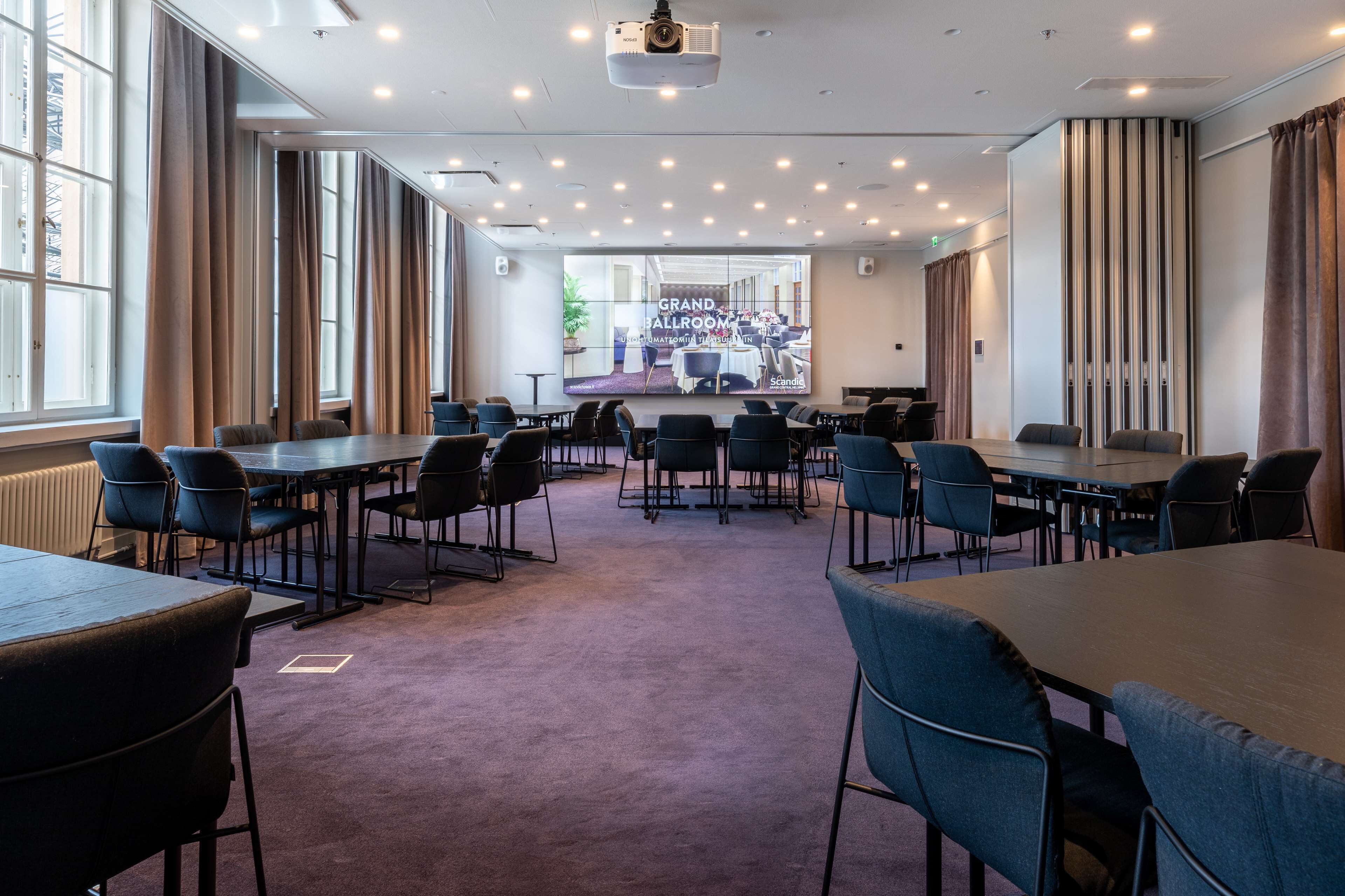 Un salón de conferencias con varias mesas y sillas, ventanas amplias y una pantalla de proyección que muestra 'Grand Ballroom'.