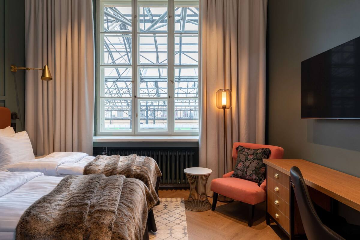 A hotel room with two beds, a desk, a chair, a TV, and a window with a view of a building structure.