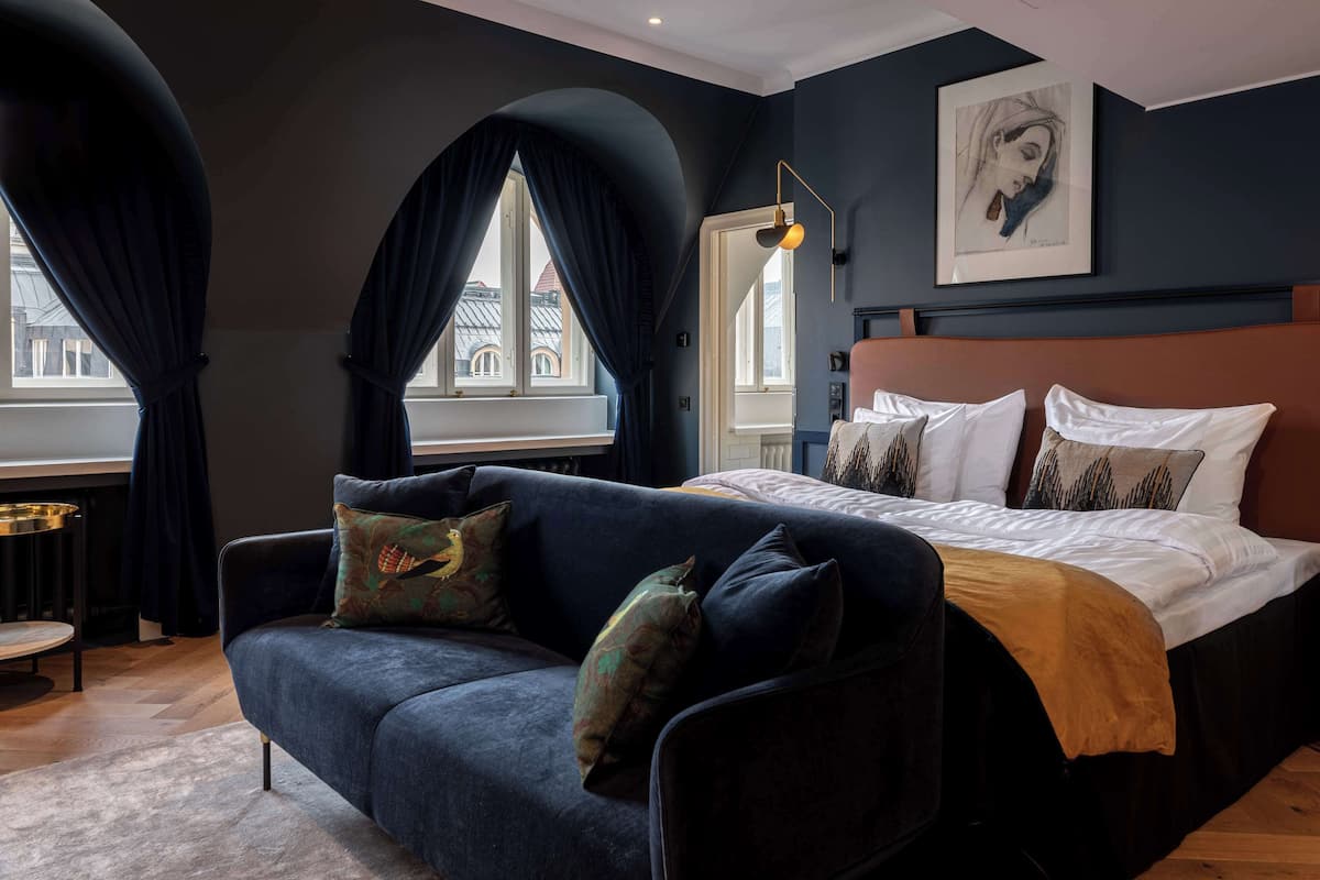 A bedroom with a dark blue sofa, a bed with white bedding, and a large window with dark curtains.