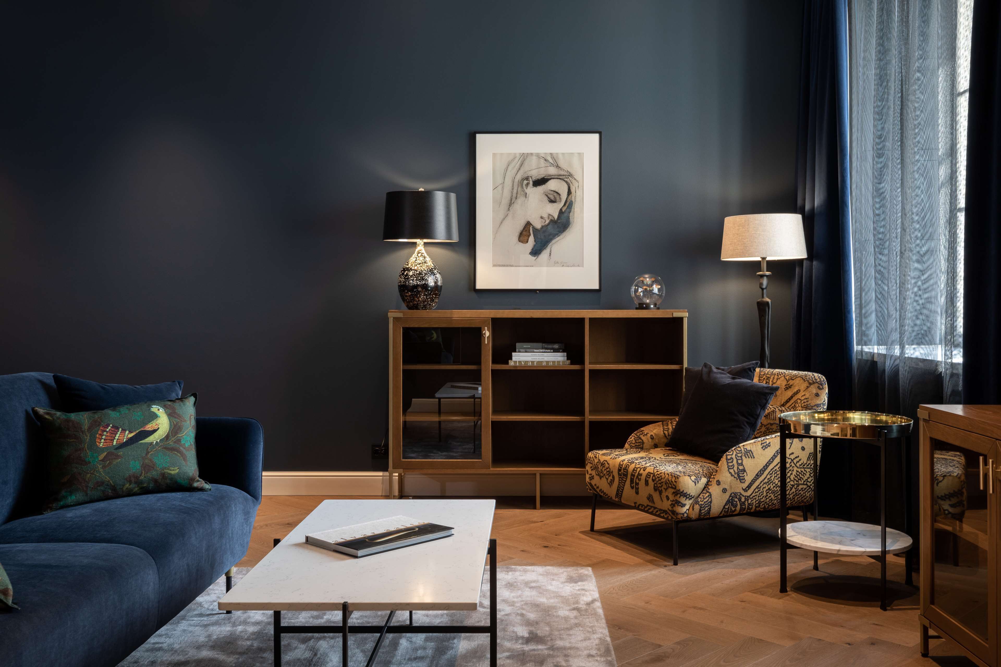 A modern living room with a dark wall, a blue sofa, a patterned armchair, a coffee table, and a bookshelf.