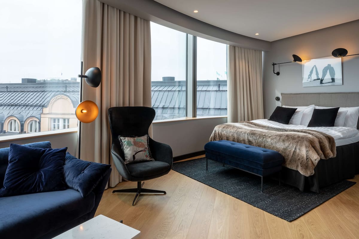 A modern hotel room with a large window, a bed with a fur throw, a dark armchair, and a blue sofa.
