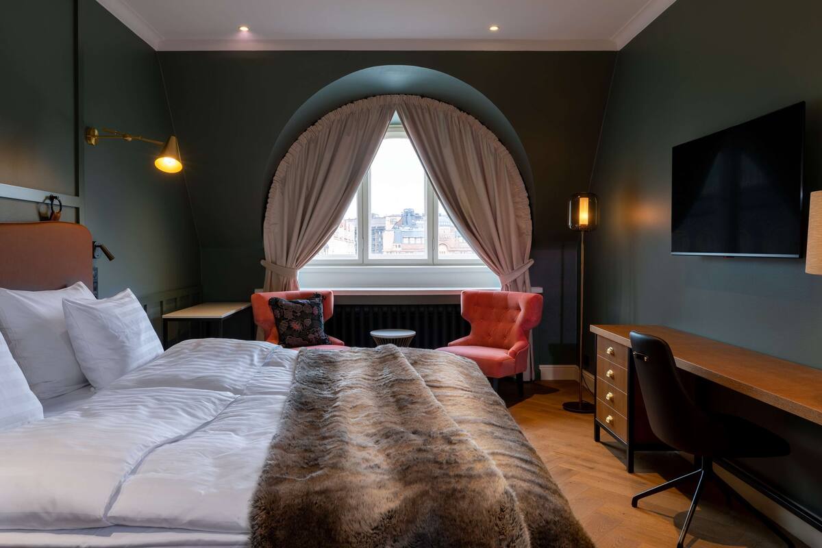 A bedroom with a bed, a desk, a chair, a window with curtains, and a TV on the wall.
