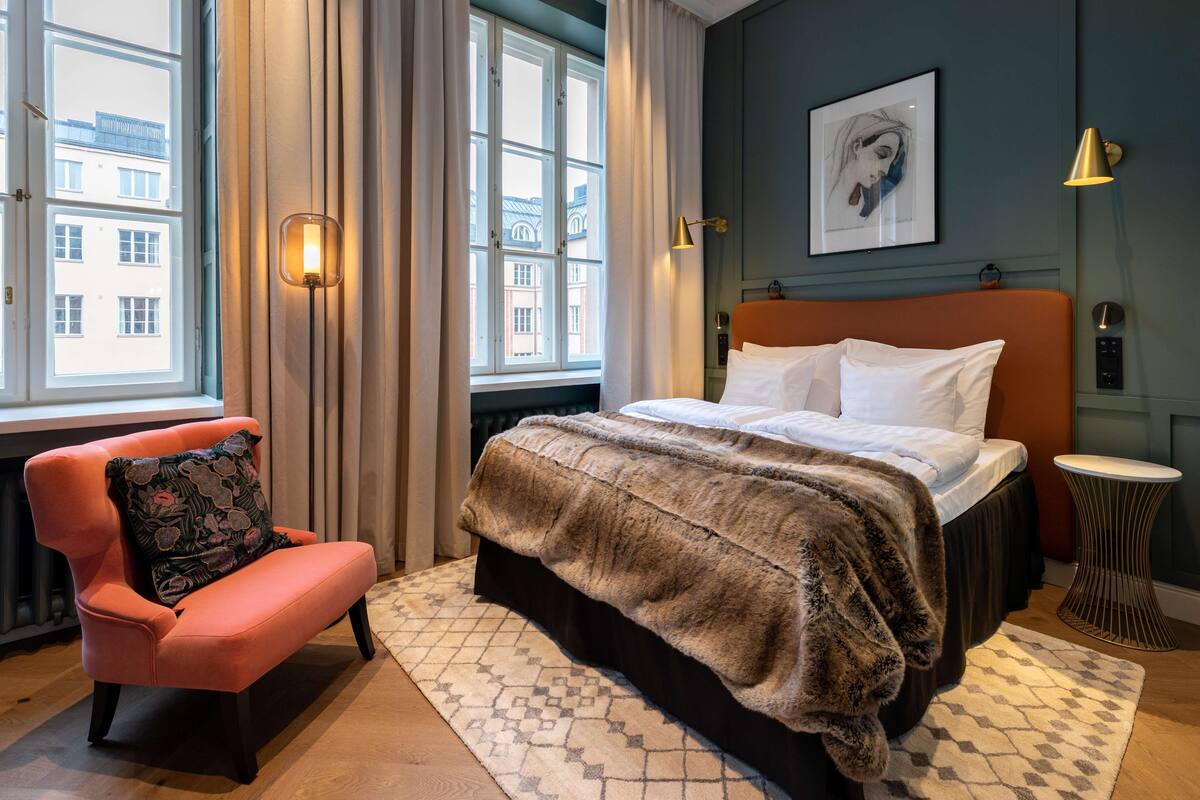 A hotel room with a bed, a chair, a window with curtains, a wall-mounted lamp, and a framed picture.