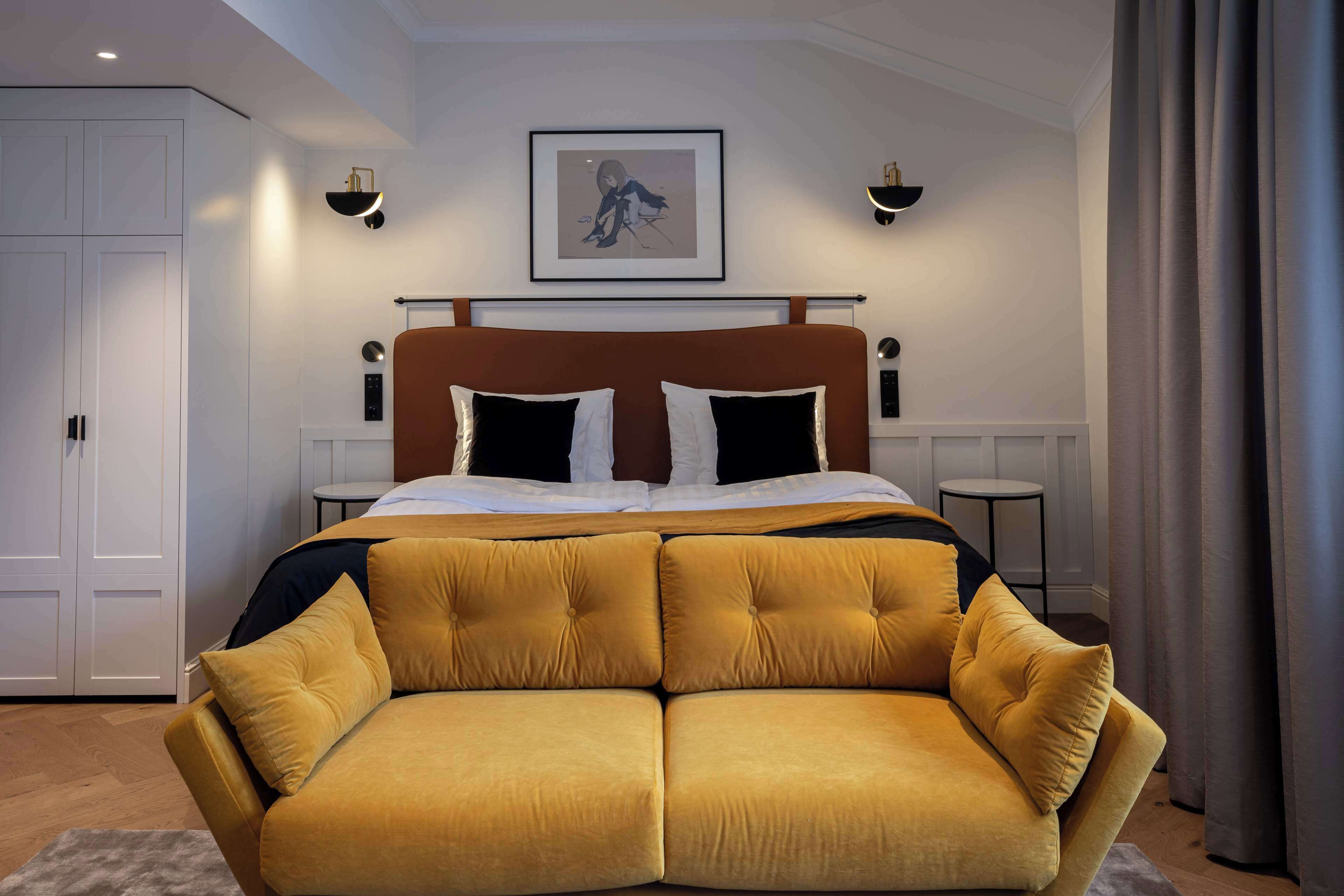 A modern bedroom with a large bed, a sofa, a framed artwork on the wall, and two wall-mounted lights.
