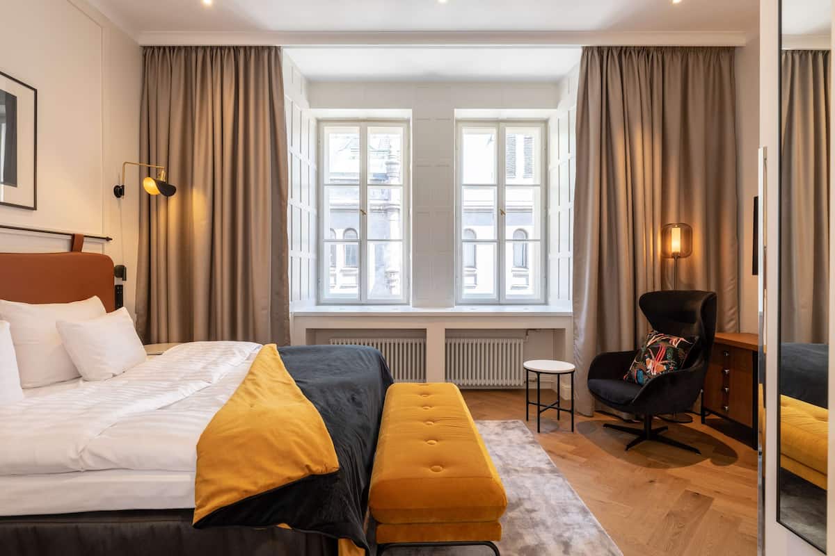 A modern hotel room with a large bed, a small side table, a chair, and a view of the city through large windows.