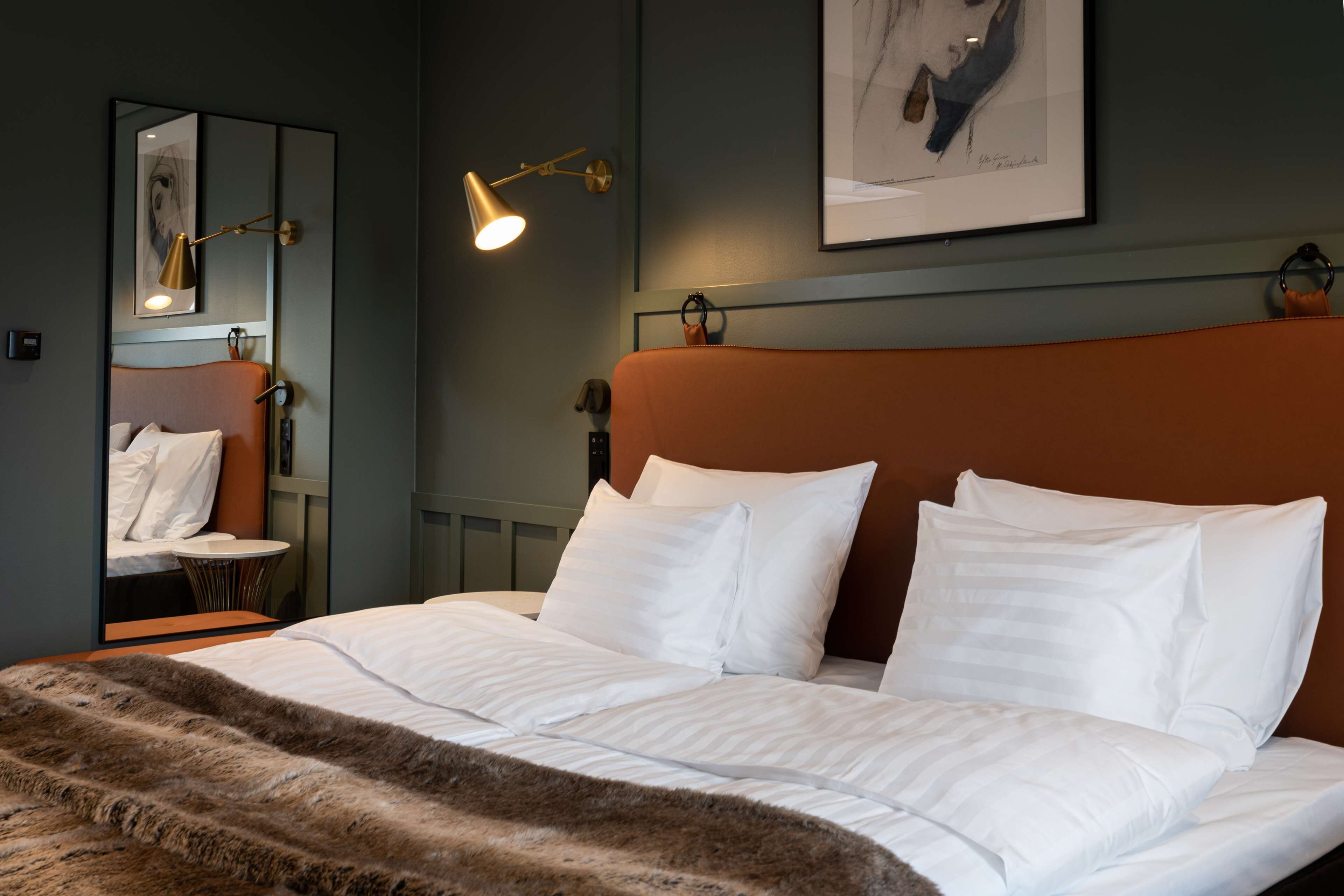 A neatly made bed with white linens and a fur throw, a bedside lamp, a framed artwork on the wall, and a small round table with a chair.