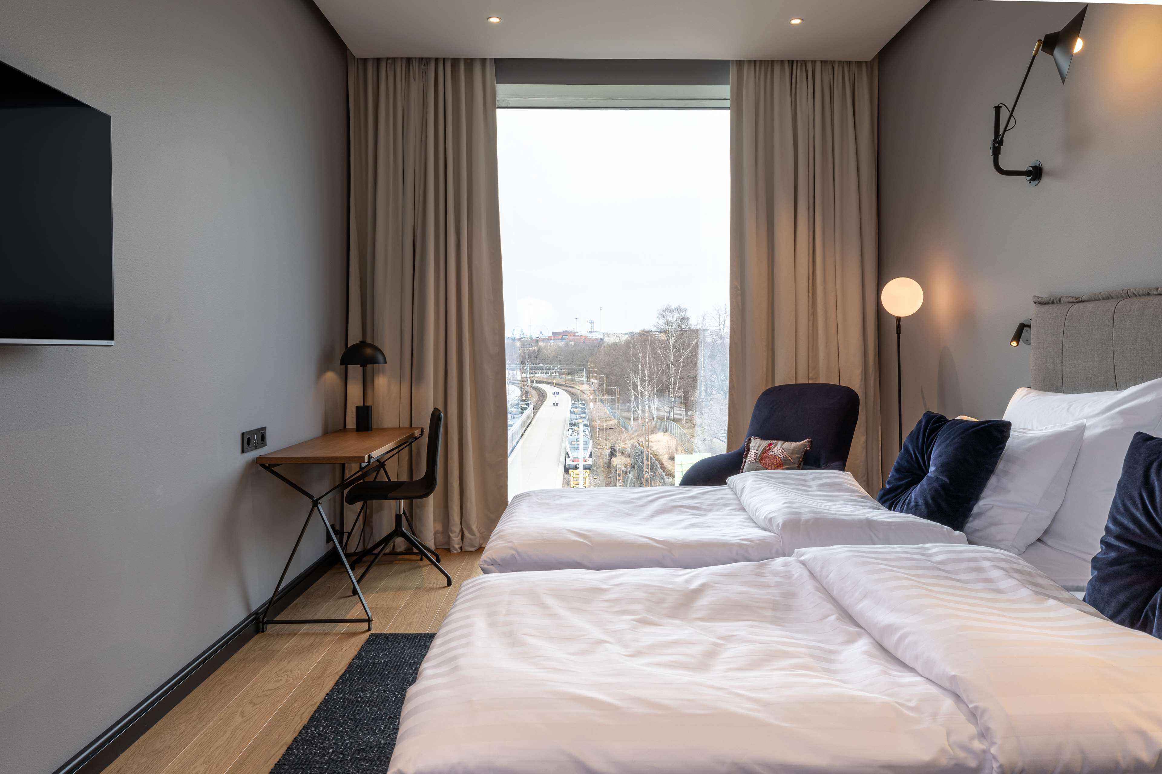 A hotel room with two beds, a desk, a chair, and a large window with a city view.