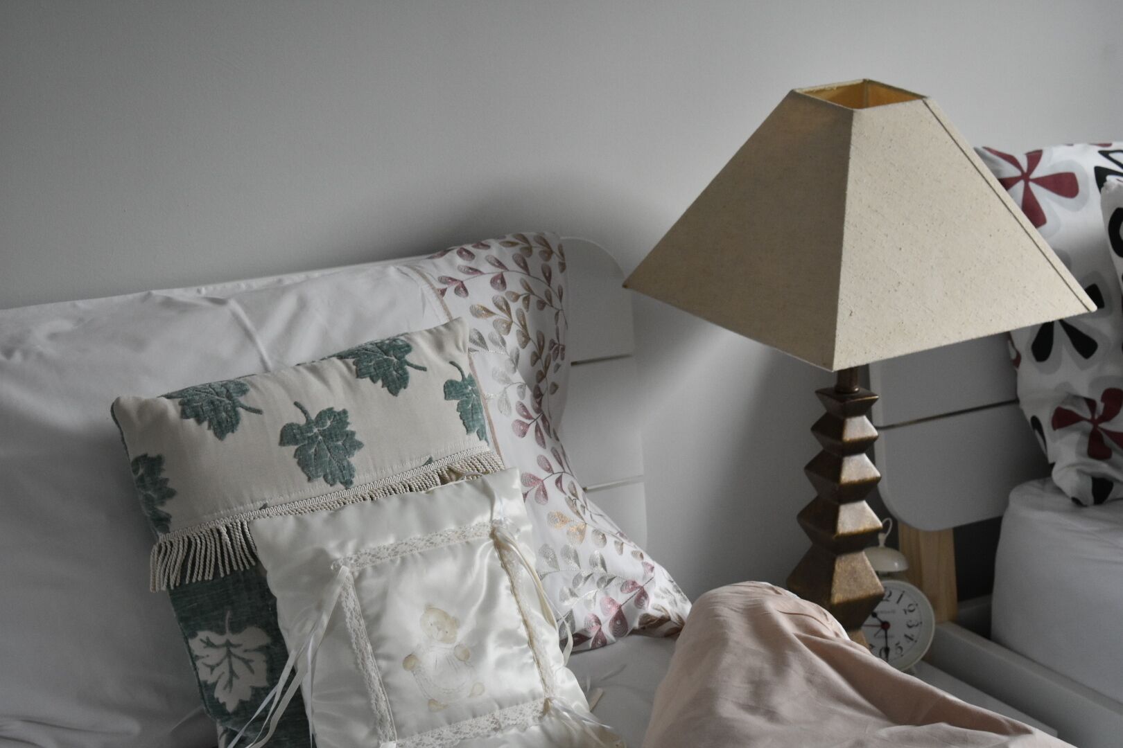 Una cama bien tendida con almohadones florales, una mesita de noche de madera con un reloj y una lámpara.