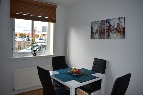 A dining area with a table set for two, a window with blinds, and a cityscape painting on the wall.