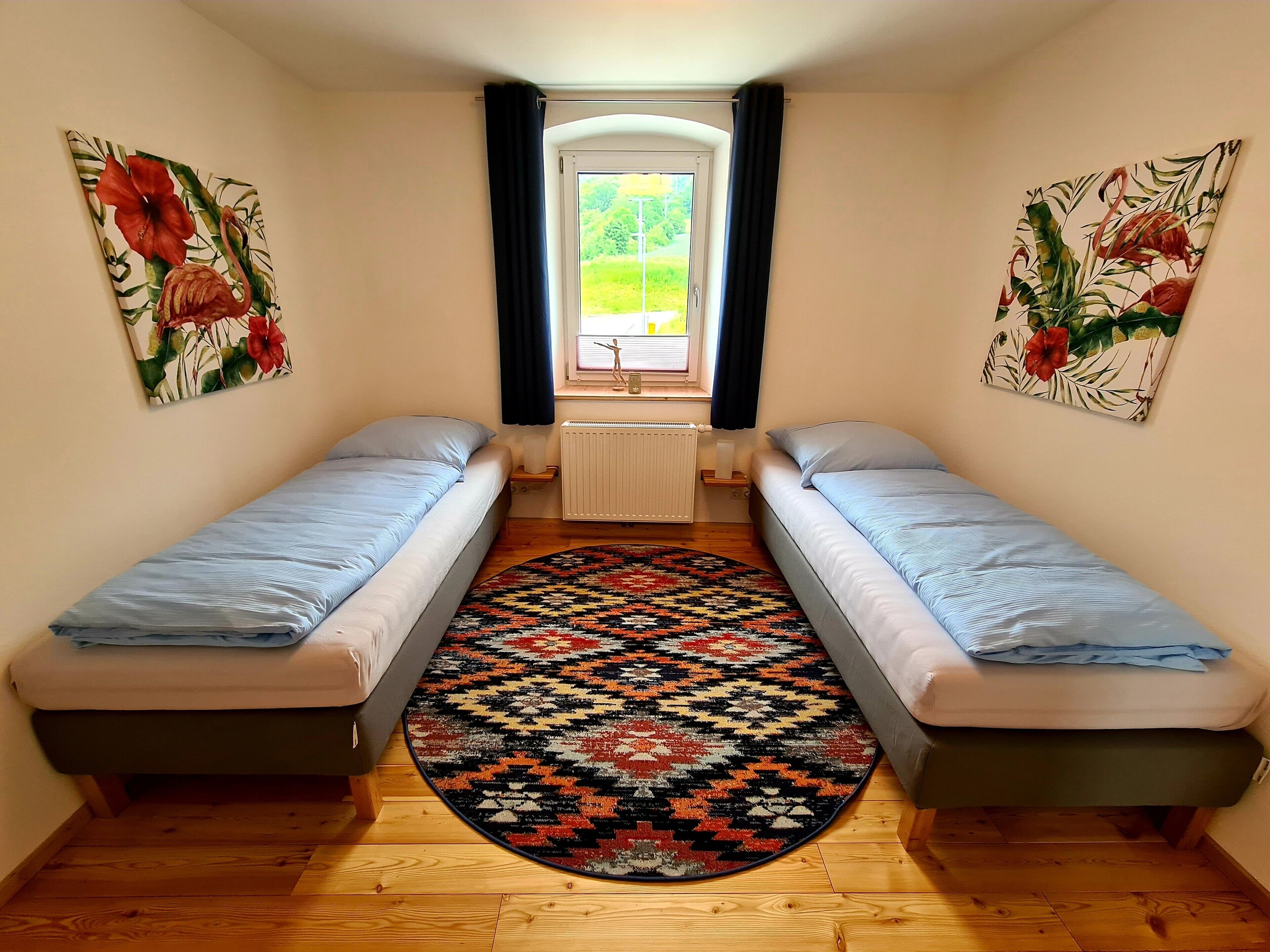 Una stanza modesta con due letti singoli, un tappeto decorato e due elementi decorativi floreali alle pareti.
