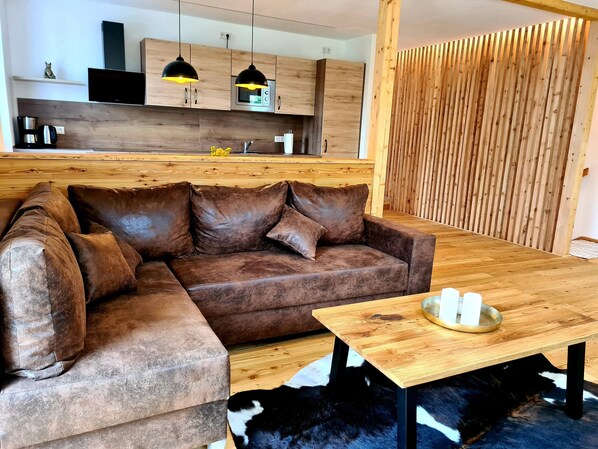 Un salon moderne avec un canapé d’angle en cuir marron, un plancher en bois, une table à manger en bois et une cuisine dotée de meubles en bois et d’un éclairage suspendu.