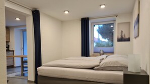 A bedroom with a bed, a bedside table, a window with curtains, a view of trees outside, and a kitchen area in the background. - Modern vacation apartment "wohnung-nummer-1" in rural area near Regensburg (Pielenhofen)