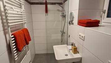 Un baño moderno con lavamanos blanco, un espejo, una ducha y un perchero con toallas rojas.