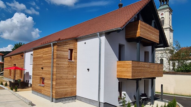 A modern building with a wooden facade, a balcony, and a clock tower in the background. - Modern vacation apartment "wohnung-nummer-1" in rural area near Regensburg (Pielenhofen)