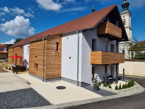Un bâtiment moderne à deux étages, doté d’une façade en bois, d’un balcon et offrant une vue sur le clocher d’une église en arrière-plan.