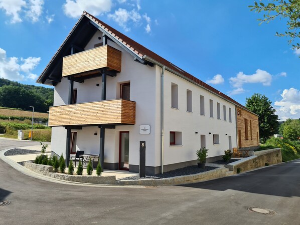 Un bâtiment moderne à deux étages, doté d’un balcon en bois, d’un petit espace extérieur aménagé en coin salon et d’une enseigne indiquant le numéro 8.