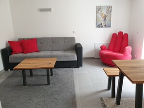 A modern living room with a grey sofa, red cushions, two wooden tables, a red chair, and a painting on the wall.