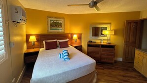 A hotel room with a bed, a ceiling fan, a nightstand with a lamp, a dresser, and a mirror. - Relaxing garden setting with ocean view A210 (Kihei)