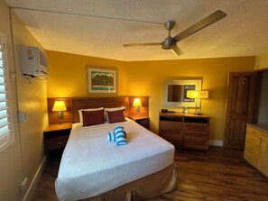 A hotel room with a bed, a ceiling fan, a nightstand with a lamp, a dresser, and a mirror.