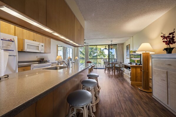 A modern kitchen with wooden cabinets, a central island with bar stools, a dining area with a view, and a large window.