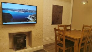 A dining area with a wooden table and chairs, a fireplace, a mounted television displaying a news channel, and a textured wall. - Immaculate 1-bed Apartment in Blackpool - Sleeps 5 (Blackpool)