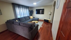 Ein Wohnzimmer mit einem Ecksofa, einem Flachbildfernseher, einer Lampe und einem kleinen Tisch.
