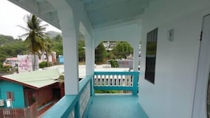 A covered balcony with a turquoise floor and white railings, overlooking a residential area with houses and a palm tree. - Veronica's - Breathe Easy - Reduit Apartment #1 (Rodney Bay)