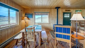 Interior de una cabaña de madera con una mesa de comedor, bancos, un sofá y vistas al exterior.