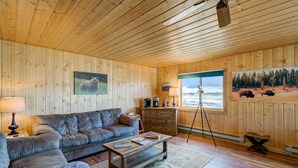 Interior acogedor de una cabaña con techo de madera, un sofá, una mesa de centro, un escritorio y un ventanal con vistas a un bosque y montañas.