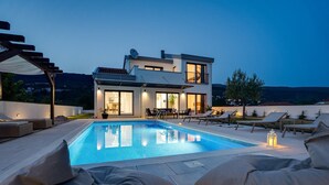 A modern two-story house with a swimming pool, outdoor seating, and a view of the hills.