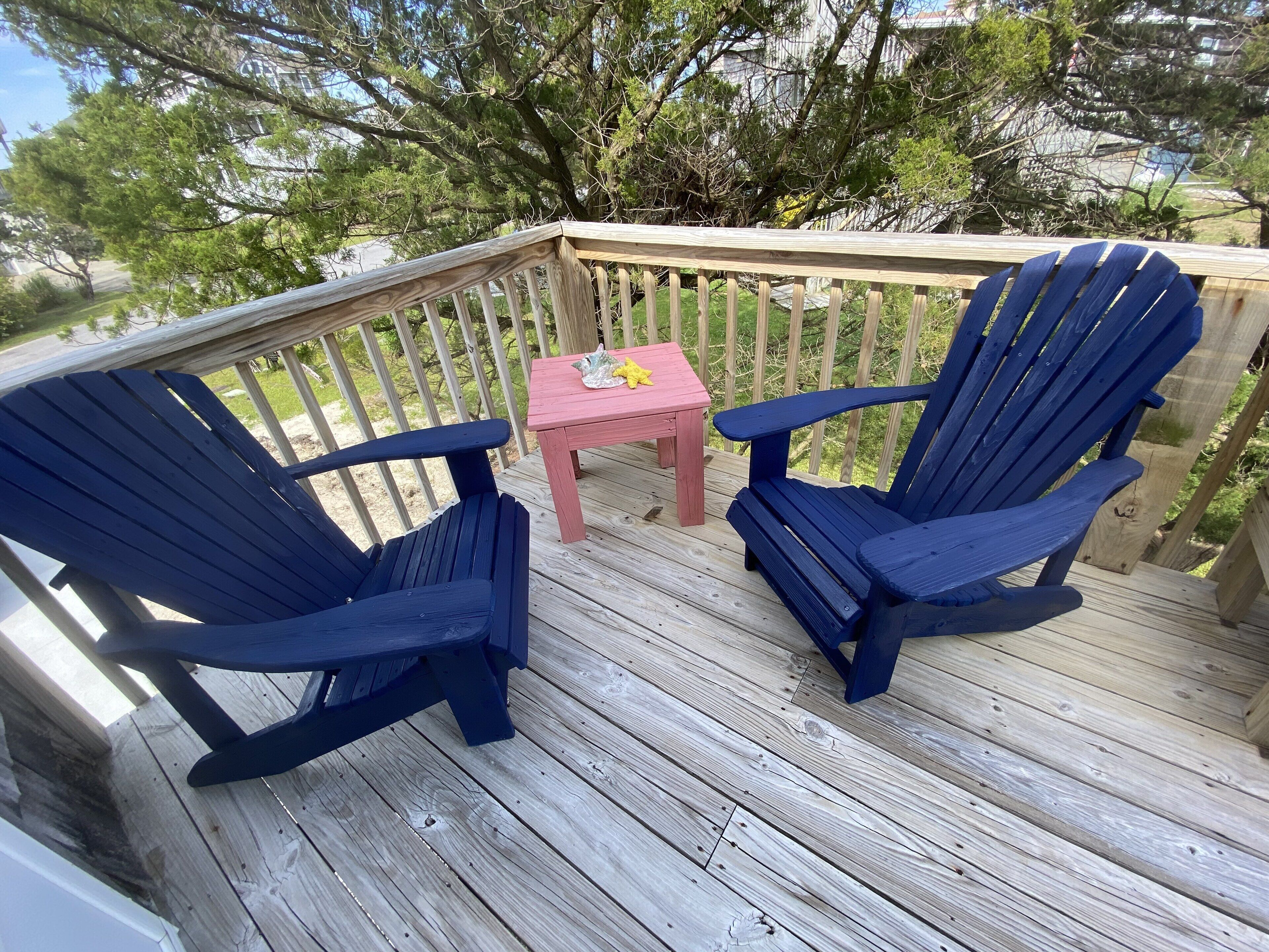 Una terraza de madera con dos sillas Adirondack azules y una mesita rosa.